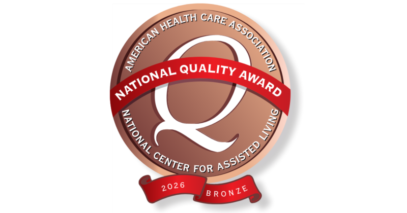 A circular bronze-colored badge with white and red accents. It features the text "American Health Care Association" around the top, "National Center for Assisted Living" around the bottom, and a red ribbon across the center with "National Quality Award" written on it. A smaller red banner at the bottom displays "2026 Bronze."